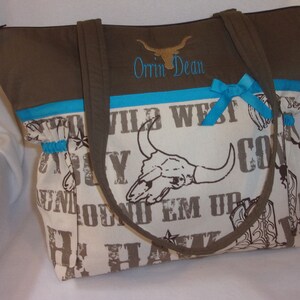 Puede incluir: Una bolsa de mano marr&oacute;n y blanca con ribete turquesa y un lazo. La bolsa tiene un dise&ntilde;o de calavera y huesos cruzados con las palabras "Wild West" y "Round Em Up". La bolsa est&aacute; personalizada con el nombre "Orin Dean".