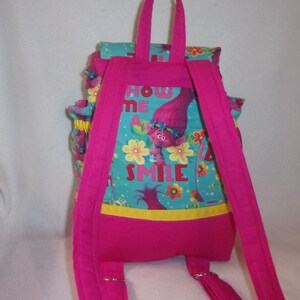 May include: Pink backpack with adjustable straps and a front pocket. The backpack features a blue and green print with a cartoon troll character and the text "Show Me A Smile".