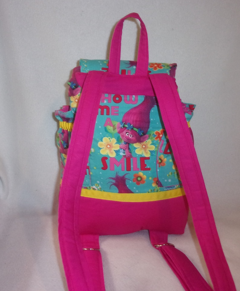 May include: Pink backpack with adjustable straps and a front pocket. The backpack features a blue and green print with a cartoon troll character and the text "Show Me A Smile".