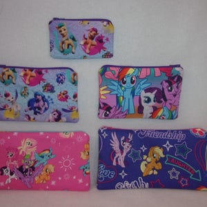 May include: Four different zippered pouches with My Little Pony characters and designs. The pouches are in various colors, including purple, blue, pink, and white. The pouches feature different characters, including Rainbow Dash, Twilight Sparkle, Pinkie Pie, and Fluttershy. The pouches have different phrases, including "Friendship is Magic", "Love", "Strength", and "Laughter".
