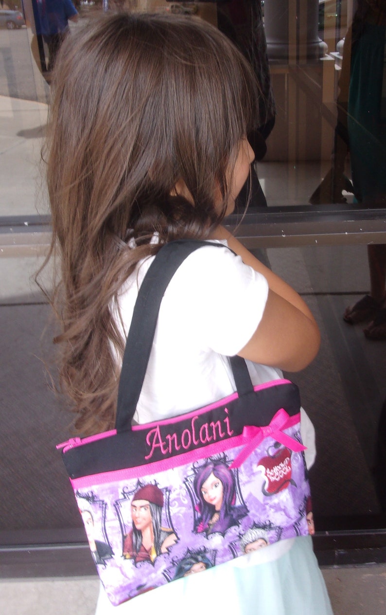 May include: A purple and pink tote bag with a black strap and the name "Anolani" embroidered on the front. The bag features a print of characters from the Disney movie "Descendants".