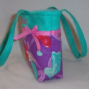 May include: Teal and purple tote bag with a pink bow and a mermaid print. The bag has a personalised name, "Olivia", embroidered on the front.