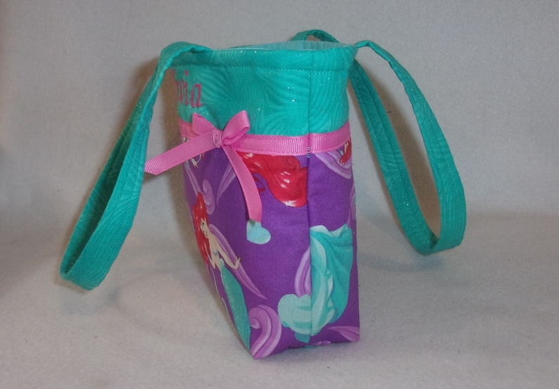 May include: Teal and purple tote bag with a pink bow and a mermaid print. The bag has a personalised name, "Olivia", embroidered on the front.
