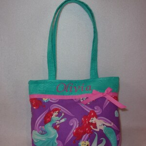 May include: A teal and purple tote bag with a pink ribbon and the name "Olivia" embroidered on the front. The bag features a repeating pattern of Ariel, the Little Mermaid, with her red hair and green tail.