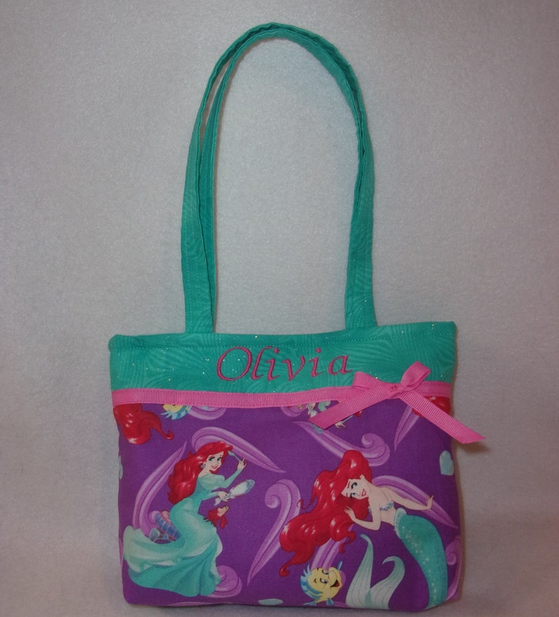 May include: A teal and purple tote bag with a pink ribbon and the name "Olivia" embroidered on the front. The bag features a repeating pattern of Ariel, the Little Mermaid, with her red hair and green tail.