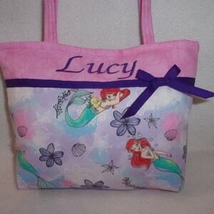 May include: Pink fabric tote bag with a purple ribbon and bow. The bag has a purple and white fabric lining with a mermaid and seashell pattern. The bag is personalised with the name "Lucy" embroidered on the front.