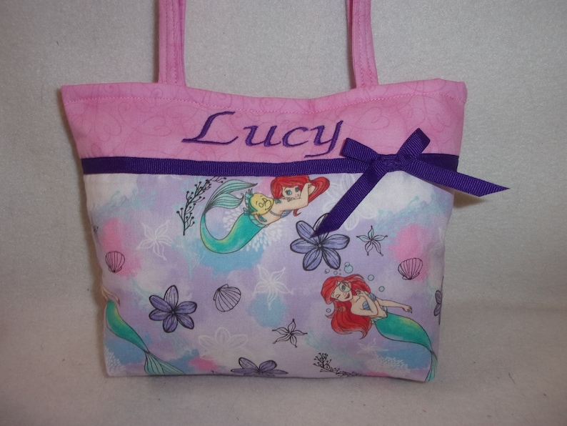 May include: Pink fabric tote bag with a purple ribbon and bow. The bag has a purple and white fabric lining with a mermaid and seashell pattern. The bag is personalised with the name "Lucy" embroidered on the front.