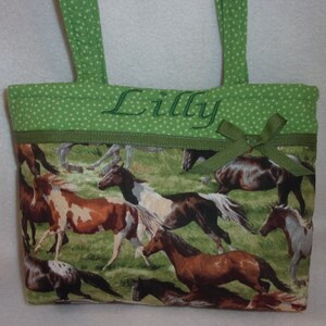 Pode incluir: Uma pequena sacola com estampa de cavalo. A sacola apresenta uma parte superior verde com bolinhas com al&ccedil;as verdes e o nome "Lilly" bordado em verde. Uma fita verde est&aacute; presa na frente.