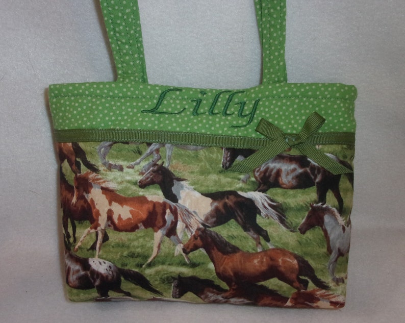 Pode incluir: Uma pequena sacola com estampa de cavalo. A sacola apresenta uma parte superior verde com bolinhas com al&ccedil;as verdes e o nome "Lilly" bordado em verde. Uma fita verde est&aacute; presa na frente.