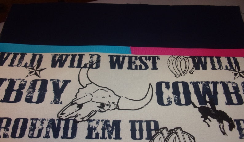 Puede incluir: Una tela blanca con un ribete azul y rosa. La tela tiene un patr&oacute;n repetido de las palabras "Wild West", "Cowboy" y "Round 'Em Up" en una fuente desgastada. El patr&oacute;n tambi&eacute;n incluye im&aacute;genes de un cr&aacute;neo de toro, una herradura y un vaquero montando un caballo.