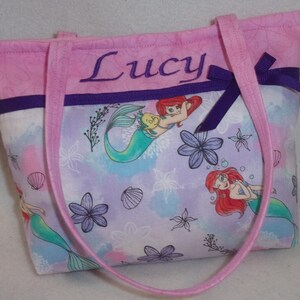 May include: Pink tote bag with purple trim and a bow. The bag is personalised with the name "Lucy" and features a mermaid print with Ariel and Flounder.
