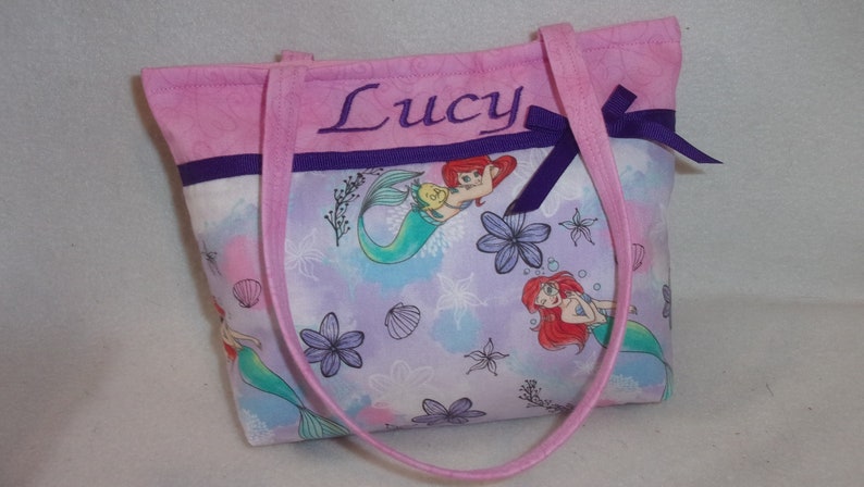 May include: Pink tote bag with purple trim and a bow. The bag is personalised with the name "Lucy" and features a mermaid print with Ariel and Flounder.