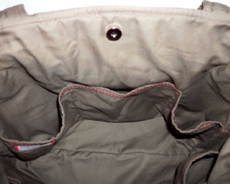 Puede incluir: El interior de una bolsa de lona marr&oacute;n con un detalle de costura roja alrededor de los bolsillos. La bolsa tiene un cierre de bot&oacute;n &uacute;nico.