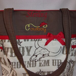 Puede incluir: Bolso de mano marr&oacute;n y blanco con una cinta roja y texto bordado que dice "Sterling Cowboy". El bolso tiene un dise&ntilde;o de calavera y huesos cruzados con las palabras "Round Em Up" impresas.