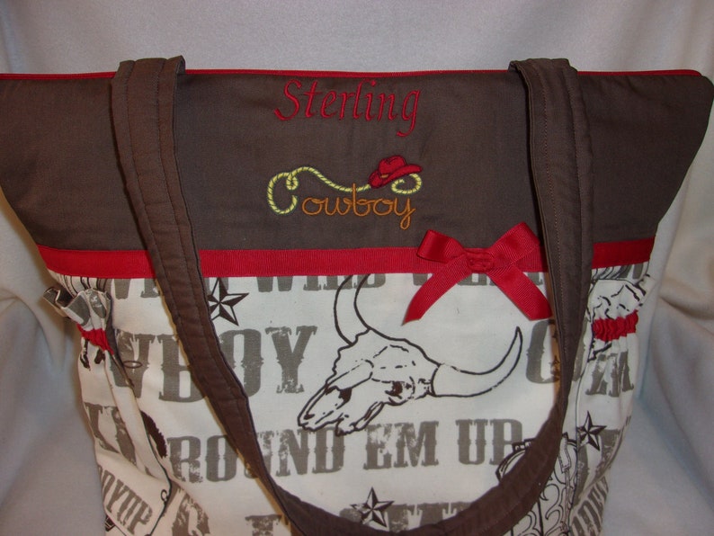 Puede incluir: Bolso de mano marr&oacute;n y blanco con una cinta roja y texto bordado que dice "Sterling Cowboy". El bolso tiene un dise&ntilde;o de calavera y huesos cruzados con las palabras "Round Em Up" impresas.