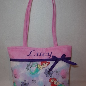 May include: Pink tote bag with a purple ribbon and a mermaid print. The bag is personalised with the name "Lucy".