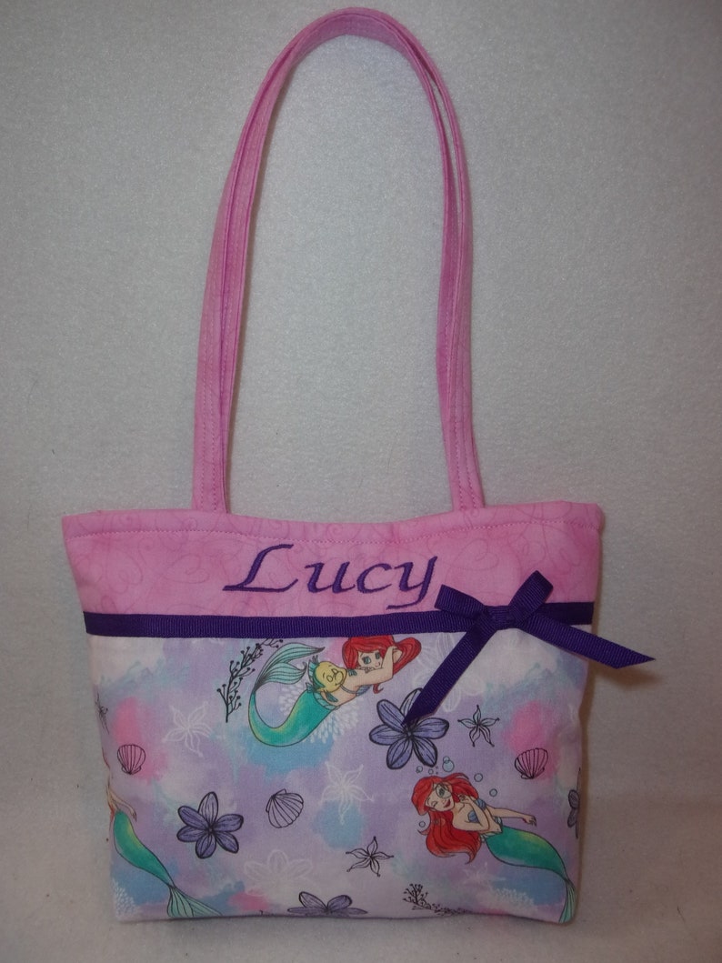 May include: Pink tote bag with a purple ribbon and a mermaid print. The bag is personalised with the name "Lucy".