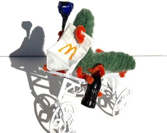 May include: A green and orange plush caterpillar riding a white tricycle, holding a blue bottle and a white paper bag with a yellow "M" logo.
