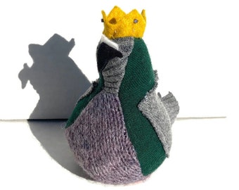 May include: A handmade fabric bird with a yellow crown. The bird is primarily purple, green, and gray, with a small pink accent. The bird is sitting on a white surface, and its shadow is visible.