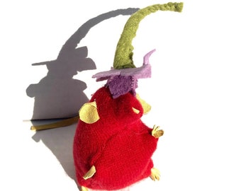 May include: A red felt mouse with a purple and green felt flower on its head. The mouse has yellow felt ears and is standing on a white surface.