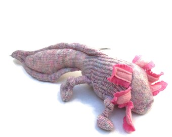May include: A knitted axolotl plush toy in shades of pink, purple, and white. The plush features detailed knitted textures, with pink frills around the head and along the sides. The toy is lying on a white surface.