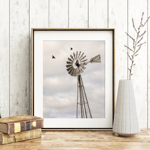 May include: A framed photograph of a vintage windmill with a cloudy sky in the background. Two birds are flying in the sky. The frame is on a wooden surface with a vase and binoculars.