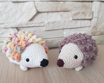 May include: Two crocheted hedgehog toys. One is white with a multi-colored spiky back and the other is white with a brown spiky back.