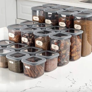 Può includere: Una collezione di contenitori trasparenti quadrati per alimenti con coperchi grigi, riempiti con varie spezie e ingredienti. Ogni contenitore ha un'etichetta. Disposti su un piano di lavoro bianco.