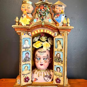 May include: A decorative, ornate cabinet with a doll head centerpiece. The cabinet is painted in gold, red, and orange with jewel accents. The doll head has blue eyes, butterfly tattoos, and a floral crown. Small portraits are displayed on the cabinet doors.