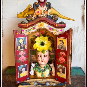 May include: A whimsical, handcrafted art piece featuring a doll head with a large yellow flower, flanked by two white cat figurines. The piece includes vintage photos, a golden dragon, and the text "no woman must limit herself to just one style."