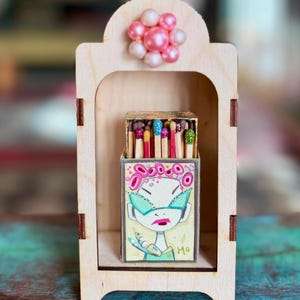May include: A wooden display case with a matchbox featuring a painted illustration of a person wearing sunglasses. The matchbox contains matches with colorful tips. Pink and white decorative beads are attached to the top of the case.