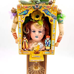 May include: A whimsical, mixed-media assemblage featuring a doll's face, miniature figures, and a birdcage. The piece is predominantly yellow with blue, green, and red accents. Decorative elements include birds, flowers, and the text "POISONOUS" and "Once upon a time".