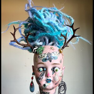 May include: A doll head sculpture with blue yarn hair, brown antlers, and insect decals. The doll has mismatched eyes, blue lipstick, and dangling earrings. The neck is adorned with a beaded collar.