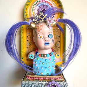 May include: A unique doll sculpture with purple hair, blue lips, and mismatched eyes. The doll wears a blue top with colorful polka dots and a beaded necklace. It is mounted on a decorative platform with a yellow and gold backdrop, adorned with embellishments.