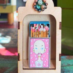 May include: A wooden display case with a blue-topped matchbox featuring a pink illustration of a person. The matchbox is filled with matches with decorative heads. A cluster of colorful beads adorns the top of the case.