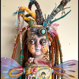 May include: A mixed media art doll with a painted face, adorned with colorful dreadlocks, a paintbrush, and decorative elements. The doll features a small pig figurine and butterfly wings. The base is a tin with a Japanese-style illustration.