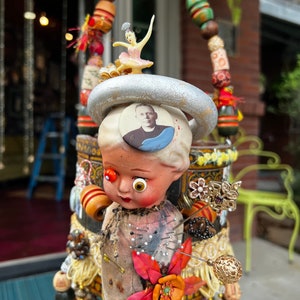 May include: A whimsical mixed media sculpture featuring a doll's head with large eyes, a flower, and a small figure of a ballerina. The sculpture is adorned with beads, fabric scraps, and other decorative elements.