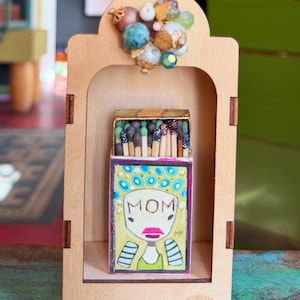 May include: A wooden display case with a matchbox featuring a hand-drawn illustration of a woman with the word "MOM". The matchbox is filled with matches. The case is adorned with decorative beads.