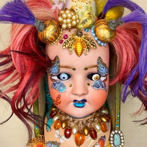 May include: A doll head sculpture with a whimsical design. The doll features red and purple hair, butterfly face paint, and a bee brooch. Adorned with a pearl necklace, gold accents, and decorative elements, it's a unique art piece.