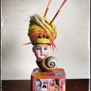 May include: A doll's head with a yellow, orange, and white yarn hairpiece sits on a wooden box. The doll's head is attached to a brown seashell. The box has a picture of a woman in a bikini on it.