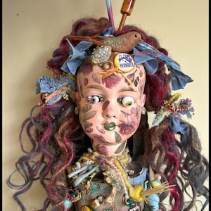 May include: A unique doll with a painted face, adorned with curly red and purple hair, blue fabric accents, and a small bird figure. The doll's face features floral designs and mismatched eyes, with a green painted mouth. The doll is wearing a decorated top.