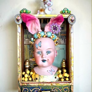 May include: A whimsical art piece featuring a doll head adorned with pink bunny ears, a jeweled crown, and butterfly tattoos. The doll is set within a decorative wooden box with a drawer, embellished with gold accents and a small ceramic bunny on top.