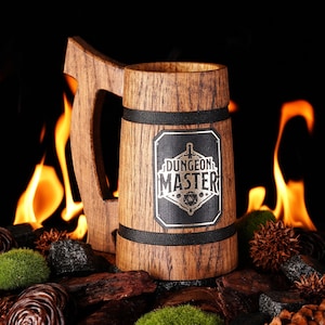 Könnte beinhalten: Ein Bierkrug aus Holz mit einem schwarzen Band und einem silbernen Aufkleber mit der Aufschrift "Dungeon Master". Der Krug steht vor einem Feuer und ist von Moos und Tannenzapfen umgeben.
