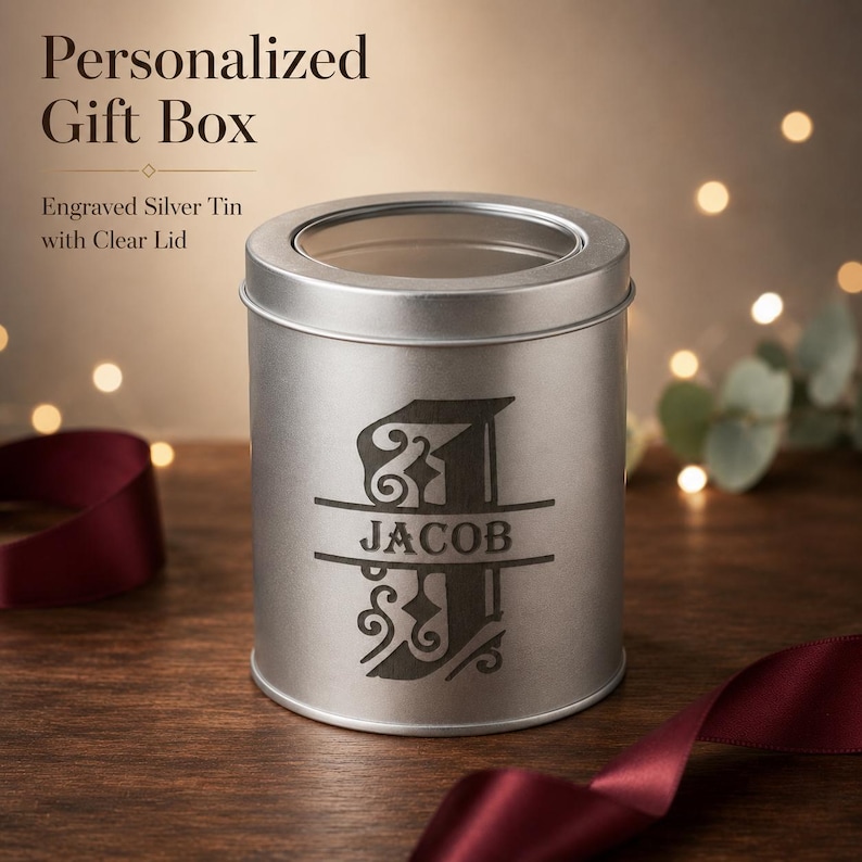 Personalized round silver metal gift box with a clear window lid and dark JACOB monogram engraving, styled with burgundy ribbon and warm lights.