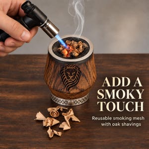 Reusable smoking mesh resting on a personalized oak whiskey cup, with oak shavings being lit by a handheld torch to create aromatic smoke.