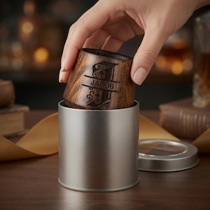 Hand placing a personalized oak shot glass inside an open silver metal gift box, showing how the wooden cup fits into the tin.