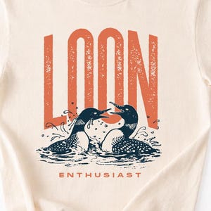 Könnte beinhalten: Cremefarbenes T-Shirt mit einer Grafik von zwei Eistauchern im Wasser. Das Wort "LOON" steht in Orange über den Eistauchern, und "ENTHUSIAST" darunter. Die Eistaucher sind blau und weiß, mit Wasserspritzern. Das T-Shirt hat einen Rundhalsausschnitt.