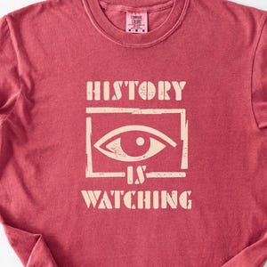 May include: A red t-shirt with the words "HISTORY IS WATCHING" in a cream-colored font. The design includes an eye within a rectangular frame. The shirt is made of a soft material and has a crew neck.