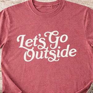 May include: A red t-shirt with the text "Let's Go Outside" in white script. The shirt is folded, with a pair of brown hiking boots and khaki pants visible. The shirt has rolled-up sleeves.