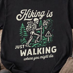 May include: Black t-shirt with a graphic of a skeleton hiker, bear, and text that reads "Hiking is just walking where you might die." The design includes green trees and a warning sign. Hiking apparel.
